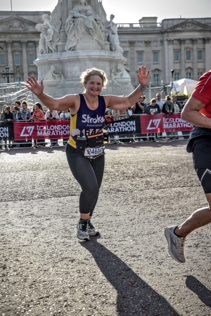 Tracy running the London Marathon - Tracy is a  woman with blonde curly hair and she's wearing a stroke assciation vest and leggings. She is smiling and waving both hands at the camera. 